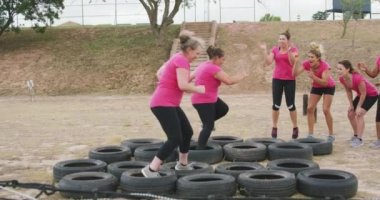 Acemi birliğinde birlikte egzersiz yapan çok ırklı, mutlu bir kadın arkadaş grubunun yan görüntüsü. İkisi lastiklerde koşuyor, diğerleri onları motive ediyor ve tezahürat yapıyor.