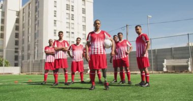 Çok ırklı erkek futbol oyuncularından oluşan bir grubun önünde soyunma takımı var, güneşin altında bir spor sahasında antrenman yapıyorlar, yan yana duruyorlar, protez bacaklı bir oyuncu elinde topla kameraya bakıyor, yavaş çekimde.