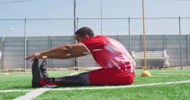 Melez bir erkek futbolcunun, protez bacaklı, takım elbiseli, güneşin altındaki bir spor sahasında antrenman yapan, esneyen, ağır çekimde