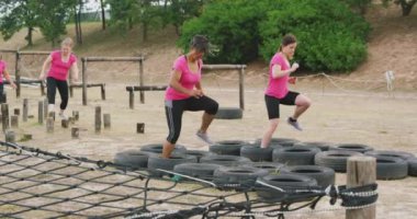 Acemi birliğinde egzersiz yapan, lastiklere basan ve arka planda ağır çekimde tahta direkler üzerinde dengede duran mutlu, beyaz ırktan pembe tişörtlü bir kadının yan görüntüsü.