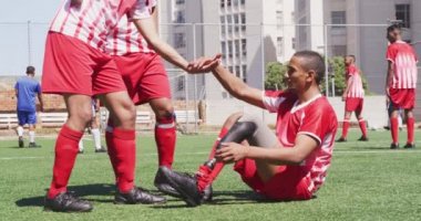 Çok ırklı takım elbiseli, güneş alan bir spor sahasında antrenman yapan erkek futbol oyuncularının yan görüntüsü, protez bacaklı engelli bir oyuncuya kolsuz ve ağır çekimde yardım ediliyor.