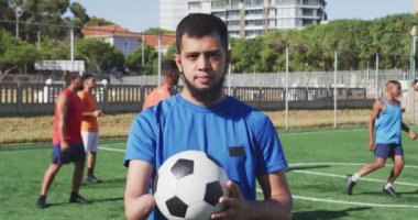 Spor kıyafetleri giyen, kameraya bakan, futbol topu tutan ve takım arkadaşlarının ağır çekimde spor sahasında antrenman yapan karışık ırktan bir erkek futbolcunun portresi.