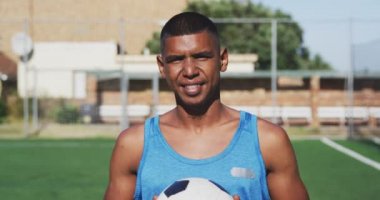 Spor kıyafetleri giyen, kameraya bakan, elinde futbol topu tutan, spor sahasında ağır çekimde duran karışık ırktan bir erkek futbolcunun portresi.
