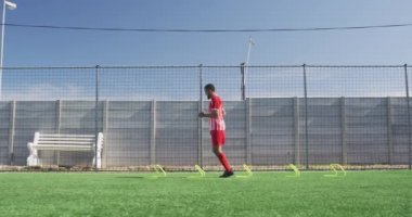 Melez bir erkek futbolcunun takım elbiseli, güneşin altında spor salonunda antrenman yapan, yavaş çekimde düşük engellerden atlayan