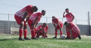 Çok ırklı erkek futbol oyuncularından oluşan bir grubun yan görüntüsü. Takım elbisesi giyiyorlar, güneşin altında spor salonunda antrenman yapıyorlar, yanlarında ağır çekimde bir topla dinleniyorlar.
