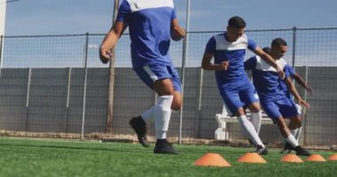 Takım elbiseli, spor sahasında antrenman yapan, ağır çekimde kozalaklara dokunup koşan çok ırklı erkek futbolcuların yan görüntüsü.