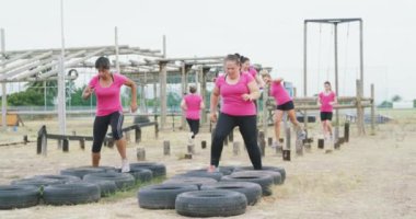 Pembe tişört giyen, acemi birliğinde egzersiz yapan, lastiklerde koşan ve ağır çekimde tahta direklerde dengede duran mutlu çoklu etnik kadın grubunun yan görüntüsü.