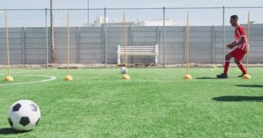 Takım elbisesi giyen, güneş alan bir spor sahasında antrenman yapan, direkler arasında ağır çekimde slalom yapan çok ırklı erkek futbolcuların yan görüntüsü.