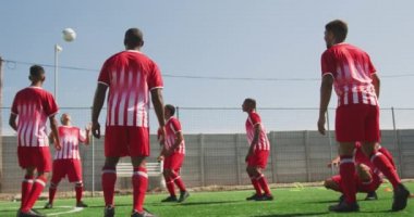 Çok ırklı erkek futbol oyuncularından oluşan bir grubun ön görüntüsü. Takım elbisesi giyiyor, güneşin altında bir spor sahasında antrenman yapıyor, yavaş çekimde topu tekmeliyor ve pas veriyor.