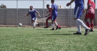 Çok ırklı iki erkek futbol takımının yan görüntüsü. Takım elbiseli, güneşin altında bir spor sahasında antrenman yapan, ağır çekimde maç yapan.
