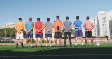 Çok ırklı erkek futbol oyuncularından oluşan bir grubun arka planda gelişigüzel spor kıyafetleri içinde güneşli bir günde bir oyun sahasında elleri arkada, yanlarında ağır çekimde bir futbol topu duruşu.