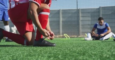 Güneşli bir günde her bir yandan oynanan futbol maçında sahada karışık ırktan erkek futbolcunun düşük kesimi arka planda takım arkadaşlarıyla ayakkabı bağları bağlıyor.