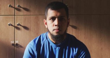 Hayal kırıklığına uğramış, spor giyimli, soyunma odasında oturan, yavaş çekimde kameraya bakan karışık ırktan bir futbolcunun portresi.