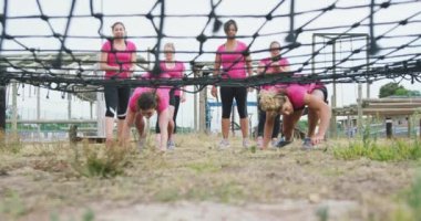 Pembe t-shirtler giyen, kampta birlikte egzersiz yapan, sıralarını bekleyen ve ağır çekimde ağların altında sürünen mutlu, çok ırklı bir kadın grubunun alçak açılı görüntüsü.