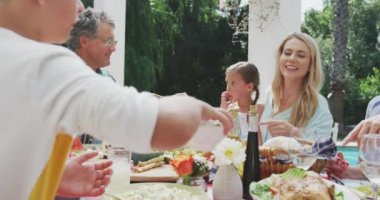 Çok nesildir birlikte iyi vakit geçiren beyaz bir ailenin yan görüntüsü. Bahçede, üzerinde yemek olan bir masanın yanında oturan bir çocuk, güneşli bir günde, ağır çekim sandviç yaparken bir yandan da bir yandan da kadınlara kase uzatıyor.