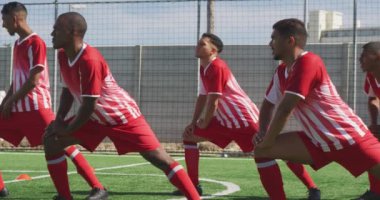 Çok ırklı bir grup erkek futbolcunun takım elbiseli, güneşin altında bir spor sahasında antrenman yapan, esneyen, ağır çekimde