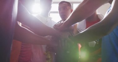 Spor giyim ve karma yarış koçu giymiş, soyunma odasında oturan, etkileşim halinde olan, yavaş çekimde el ele tutuşan çok ırklı erkek futbol oyuncularının ön görüntüsü.