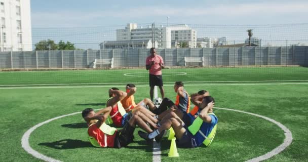 スポーツウェアと色のビブを身に着けている男性サッカー選手の多民族グループの側面図 太陽の下でスポーツフィールドでのトレーニング 行の上に座ると 彼らのコーチは 背景に立って スローモーションで — ストック動画