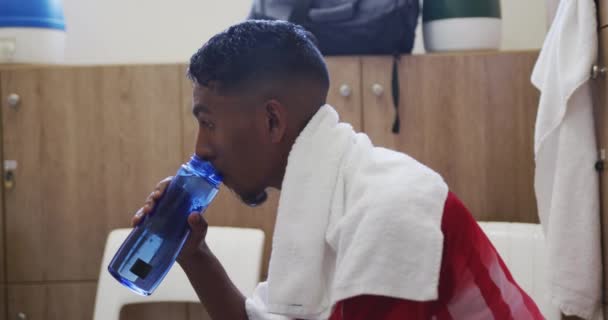 一名身穿队服 坐在更衣室里 肩上披着毛巾 慢吞吞地喝水的混合型男子足球运动员的侧影 — 图库视频影像