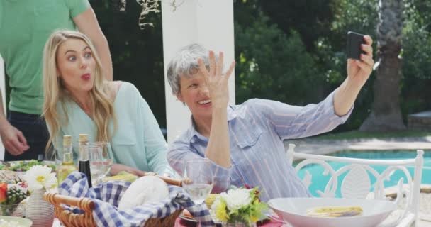 一緒に楽しい時間を過ごしている多世代の白人家族の近くのサイドビュー 庭に座って 食べ物のあるテーブルによって 年配の女性は彼女の家族と一緒に自撮りをして 晴れた日に面白い顔をしています — ストック動画