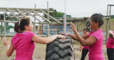 Kafkasyalı mutlu bir kadın ve kampta birlikte egzersiz yapmaktan hoşlanan melez bir kız arkadaşın yan görüntüsü, traktör lastiği ve beşlik çakma, arka planda diğer katılımcılar yavaş çekimde,