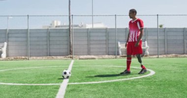 Melez bir erkek futbolcunun, protez bacaklı, takım elbiseli, güneşin altındaki bir spor sahasında antrenman yapan, topa tekme atan, ağır çekimde