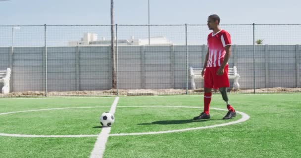 Oldalnézet egy vegyes fajú férfi focista egy műláb visel egy csapat szalag, edzés egy sportpályán a napon, rugdossa a labdát, lassított felvételen