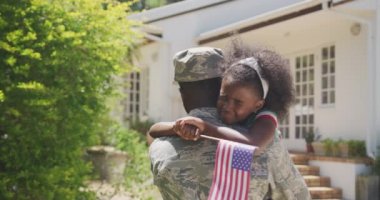 Afrika kökenli Amerikalı bir adamın bahçede, ailesiyle vakit geçirmesini, askeri üniforma giymesini, ağlayan Afrikalı Amerikalı kızını elinde bayrak tutmasını, güneşli bir günde, ağır çekimde kucaklamasını.