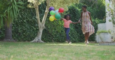 Afrika kökenli Amerikalı bir kadının kızıyla bahçede iyi vakit geçirmesi ön planda, koşuyor, zıplıyor, el ele tutuşuyor, bir kız balon tutuyor, güneşli bir günde, ağır çekimde.