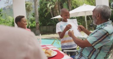 Çoklu etnik çeşitlilikte bir ailenin birlikte iyi vakit geçirmesini, bahçesinde yemek olan bir masada oturmasını, büyükbabasına bir kağıt parçası göstermesini, güneşli bir günde herkesin ona tezahürat etmesini.