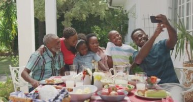 Çok nesilli çok ırklı bir ailenin birlikte iyi vakit geçirmesinin yan görüntüsü. Bahçede, üzerinde yemek olan bir masada oturan, akıllı telefon tutan, güneşli bir günde ailesiyle selfie çeken yetişkin bir adam.