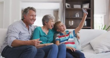 Üst düzey beyaz bir çiftin dairede, koltukta otururken, torunlarıyla birlikte akıllı bir telefon tutarken, birlikte selfie çekerken, komik suratlar yaparken, ağır çekimde...