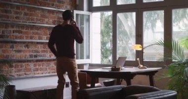 Arka tarafta, kısa siyah saçlı, evde çalışan, akıllı telefondan konuşan ve pencereye bakan melez bir adam var..