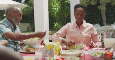 Afrika kökenli Amerikalı bir kadın ve kıdemli bir Afro-Amerikan erkeği, güneşli bir günde, bir bahçede, bir masada oturup, tabaklarına yemek koyarken, birlikte iyi vakit geçiriyorlar.