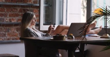Uzun sarı saçlı, çekici beyaz bir kadının oturma odasında ayaklarını masaya dayayıp bilgisayarını kullanarak ve akıllı telefonuyla mesajlaşarak evde vakit geçirmesinin yan görüntüsü..
