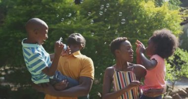 Afrika kökenli Amerikalı bir ailenin bahçede iyi vakit geçirmesini, ayakta durup kabarcıklar çıkarmasını, güneşli bir günde, yavaş çekimde ebeveynlerin çocuklarını havada tutmasını.