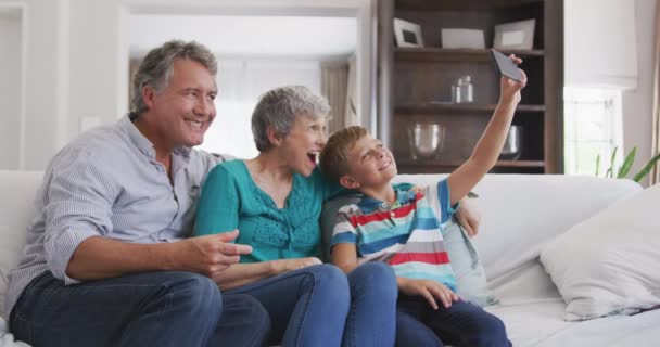 一对年长的白人夫妇在公寓里享受时光 坐在沙发上 孙子拿着智能手机 一起自私自利 慢吞吞地做鬼脸 — 图库视频影像