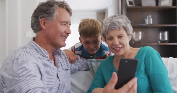 一对年长的白人夫妇坐在公寓里享受时光 坐在沙发上 用智能手机 身后站着他们的孙子 动作缓慢 — 图库视频影像