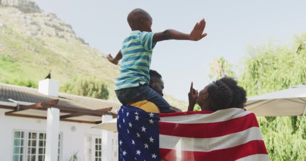 在一个阳光灿烂的日子里 父母们慢吞吞地抱着他们的孩子 带着他们在花园里玩得很开心 站在那里 裹着美国国旗 — 图库视频影像