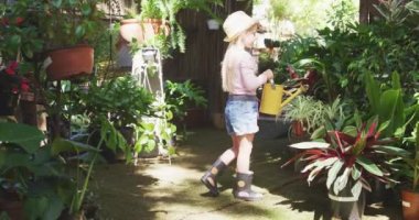 Beyaz tenli, uzun sarı saçlı, güneşli bir bahçede eğlenen, çiçekleri sulama tenekesiyle sulayan, Wellingtons giyen ve şapkasını yavaşça takan kızın yan görüntüsü.