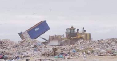 Aracın üzerinde uçan ve çöpleri temizleyen kuş sürüsü arka planda bulutlu bulutlu gökyüzü olan çöplerle dolu bir çöp yığını üzerinde ağır çekimde çalışıyor.
