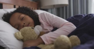 Karışık ırk kızlarının yan görünüşü evde eğleniyor, yatakta uzanıyor, uyuyor, gülümsüyor, oyuncak ayıya sarılıyor, ağır çekimde