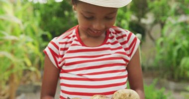 Ön planda melez bir kız var. Bahçede vakit geçiriyor. Ağır çekimde taze soyulmuş patatesleri sunuyor.