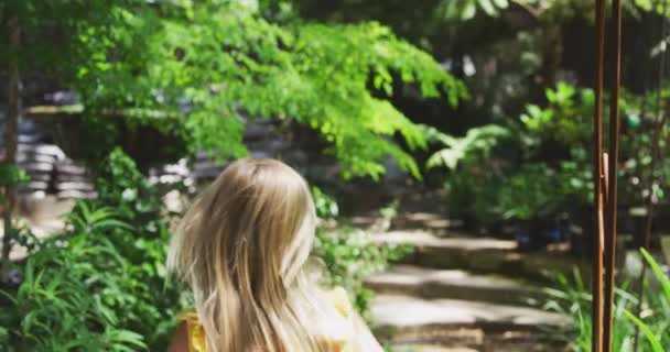 在一个阳光灿烂的花园里 一个有着长长的金发的白人女孩兴奋地沿着小路奔跑着 慢吞吞地看着她的背影 — 图库视频影像