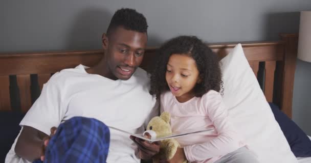 一个非洲裔美国人和他的混血儿在一起享受家里的时光 坐在床上 一个男人在看他的女儿看书 慢吞吞地笑着 — 图库视频影像