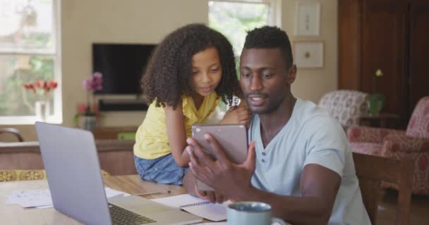 一个非裔美国人和他的混血女儿在家里享受着时光 坐在桌子旁边 一个男人拿着一个数码平板电脑 带着微笑 慢动作地一起观看着 — 图库视频影像