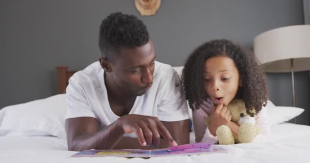 アフリカ系アメリカ人の男と彼の混合人種の娘の家で一緒に時間を楽しんで ベッドの上に横たわって 男はゆっくりと動きながら 彼の娘に本を読んでいます — ストック動画