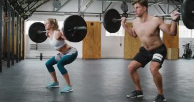 Spor kıyafetleri giyen beyaz bir erkek ve kadının spor salonunda antreman yaparken, ayakta ve ağırlık antrenmanı yaparken, halterlerini omuzlarında tutarken ve yükselirken yan görüntüsü. Adam üstsüz.