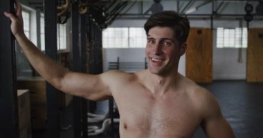 Mutlu, üstsüz, yakışıklı, atletik, beyaz bir adamın portresi spor salonunda çapraz antrenman yapıyor, mola veriyor, kameraya bakıyor ve gülümsüyor.