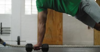 Yan görüntüde spor kıyafetleri giyen beyaz bir adam spor salonunda çapraz antrenman yapıyor, halterlerle ağırlık çalışması yapıyor, dambılları tutarak baskı pozisyonunda yatıyor ve yedek ağırlık kaldırıyor.
